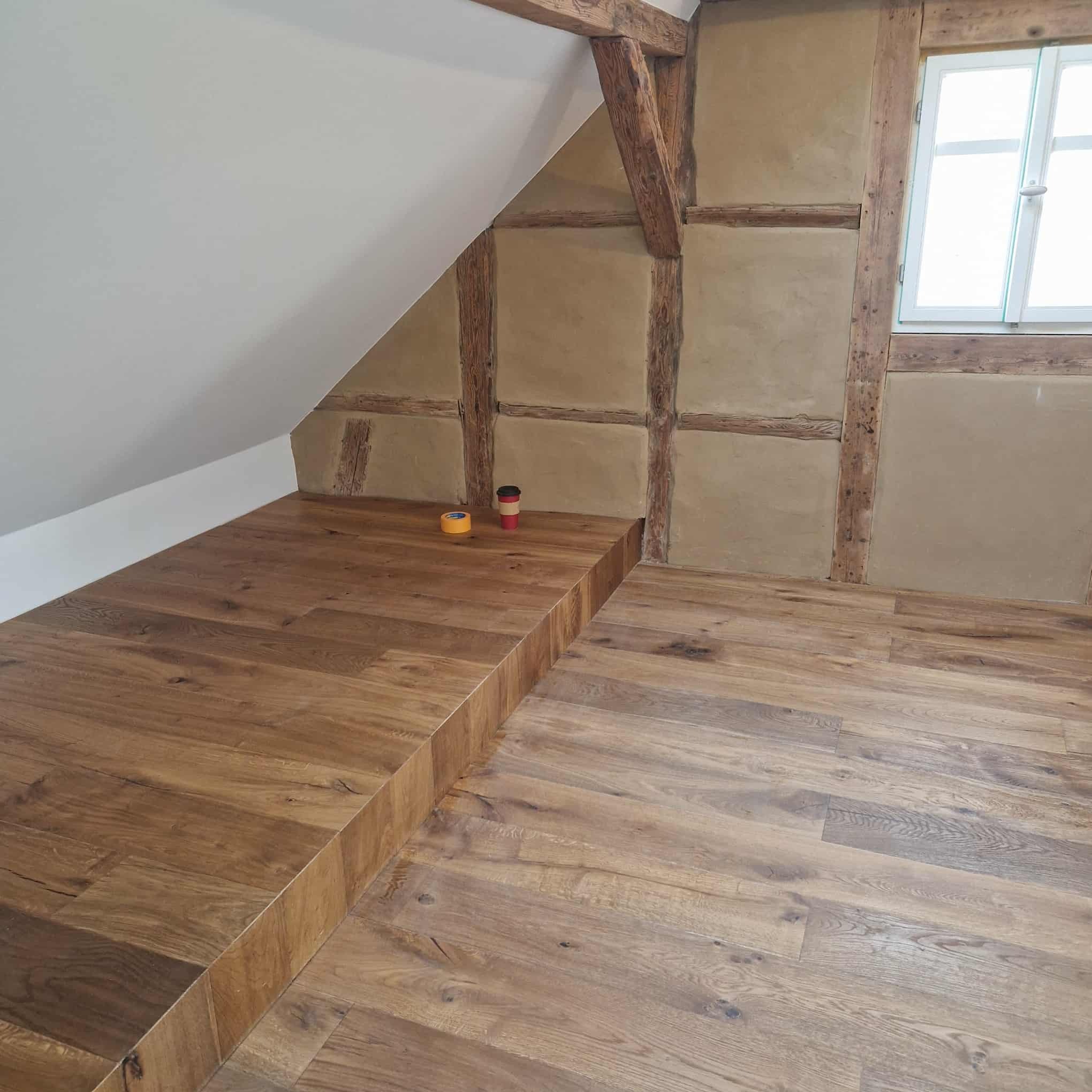 Podłoga z desek w odcieniach brązu w pokoju na poddaszu z drewnianą konstrukcją ścian i oknem. Widoczne detale wykończenia i naturalne światło.