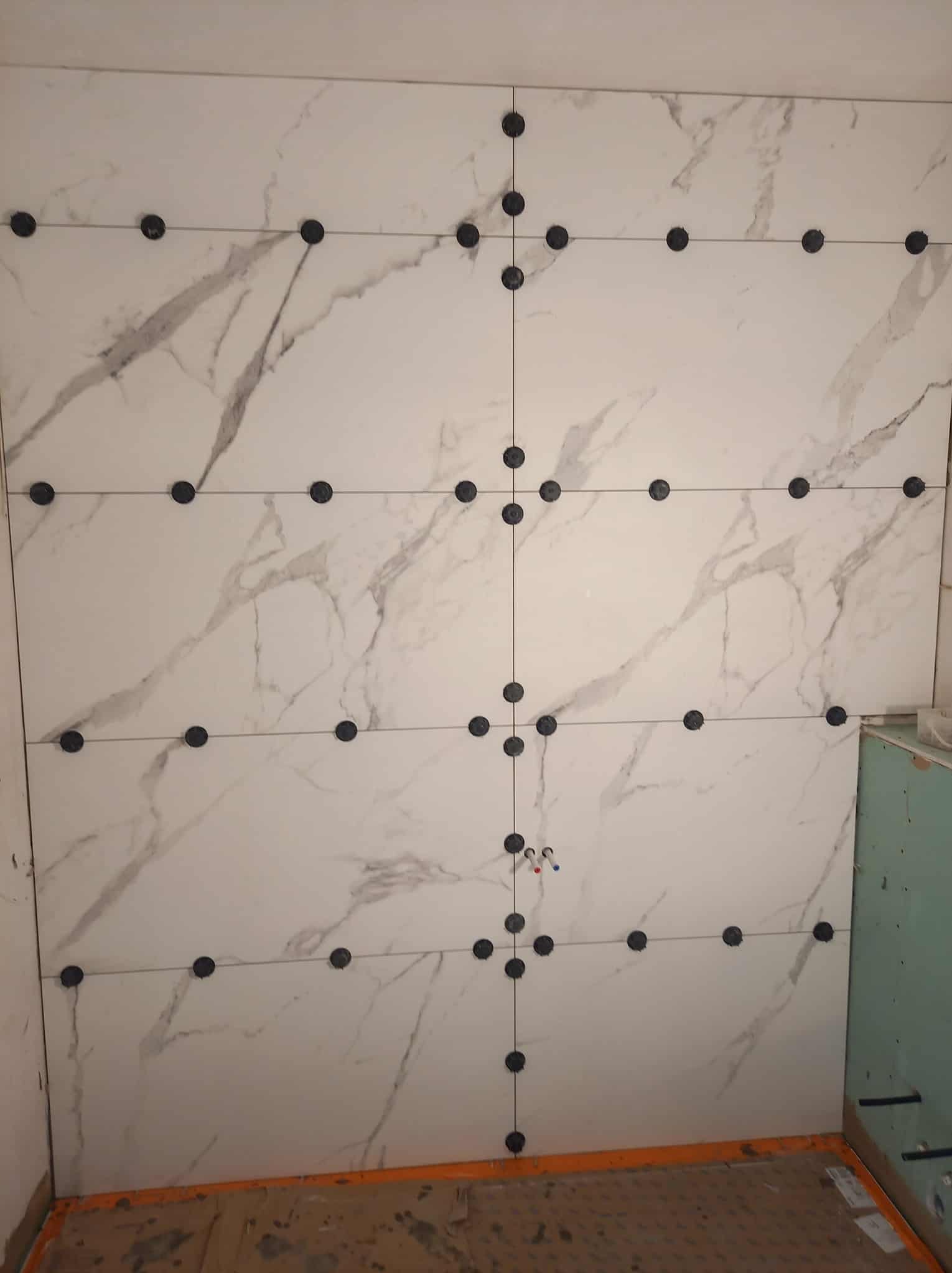 Ściana z białych płytek imitujących marmur, z czarnymi krzyżykami dystansowymi, w trakcie układania. Widoczne fragmenty podłogi i sąsiednich ścian.