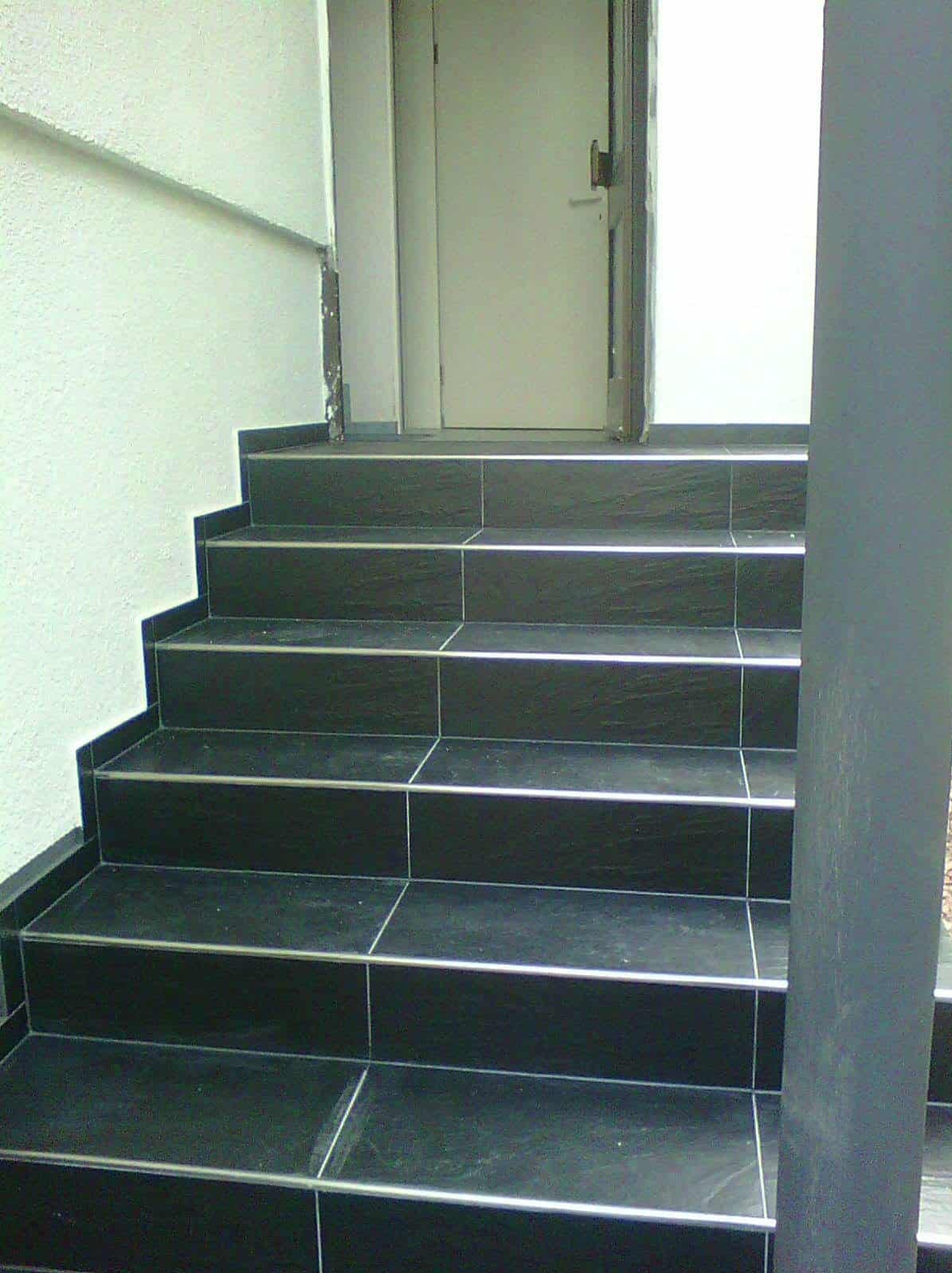 Wykończone schody zewnętrzne z ciemnych płytek z metalowymi listwami, prowadzące do drzwi wejściowych. Widoczne fragmenty ściany i słupa.