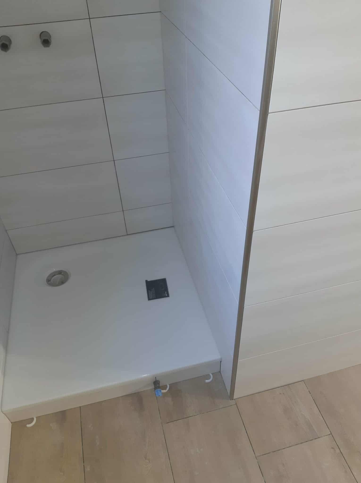 Nowoczesny brodzik z odpływem liniowym i białymi, prostokątnymi płytkami ściennymi, wykończony listwą. Podłoga wyłożona płytkami imitującymi drewno.