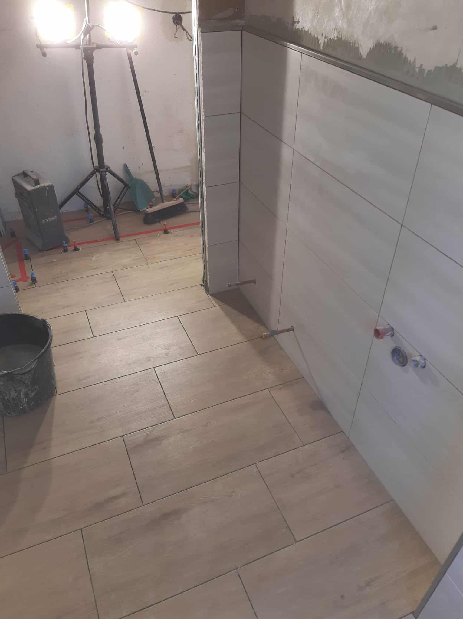 Układanie płytek w łazience: beżowe płytki podłogowe imitujące drewno, białe płytki ścienne z poziomymi paskami, widoczne narzędzia i oświetlenie budowlane.