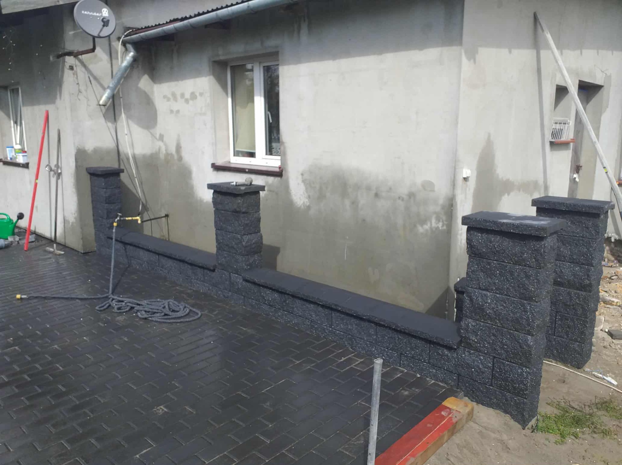 Nowoczesny murek z ciemnych, grafitowych bloczków, wykończony gładkimi płytami. Widoczna kostka brukowa i fragment elewacji budynku z oknem. Wąż ogrodowy na pierwszym planie.