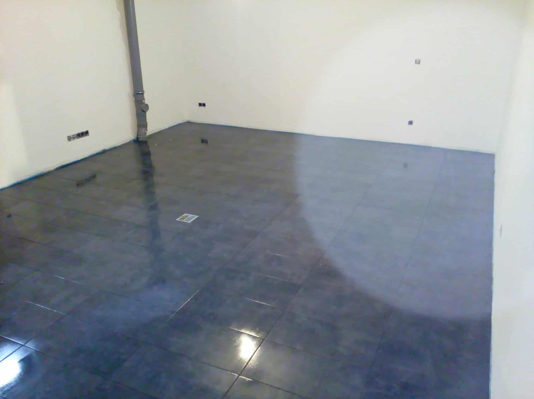 Błyszcząca, ciemna podłoga z płytek w pustym pomieszczeniu z białymi ścianami. Widoczny odpływ podłogowy i rura przy ścianie. Świeżo położone płytki podłogowe.