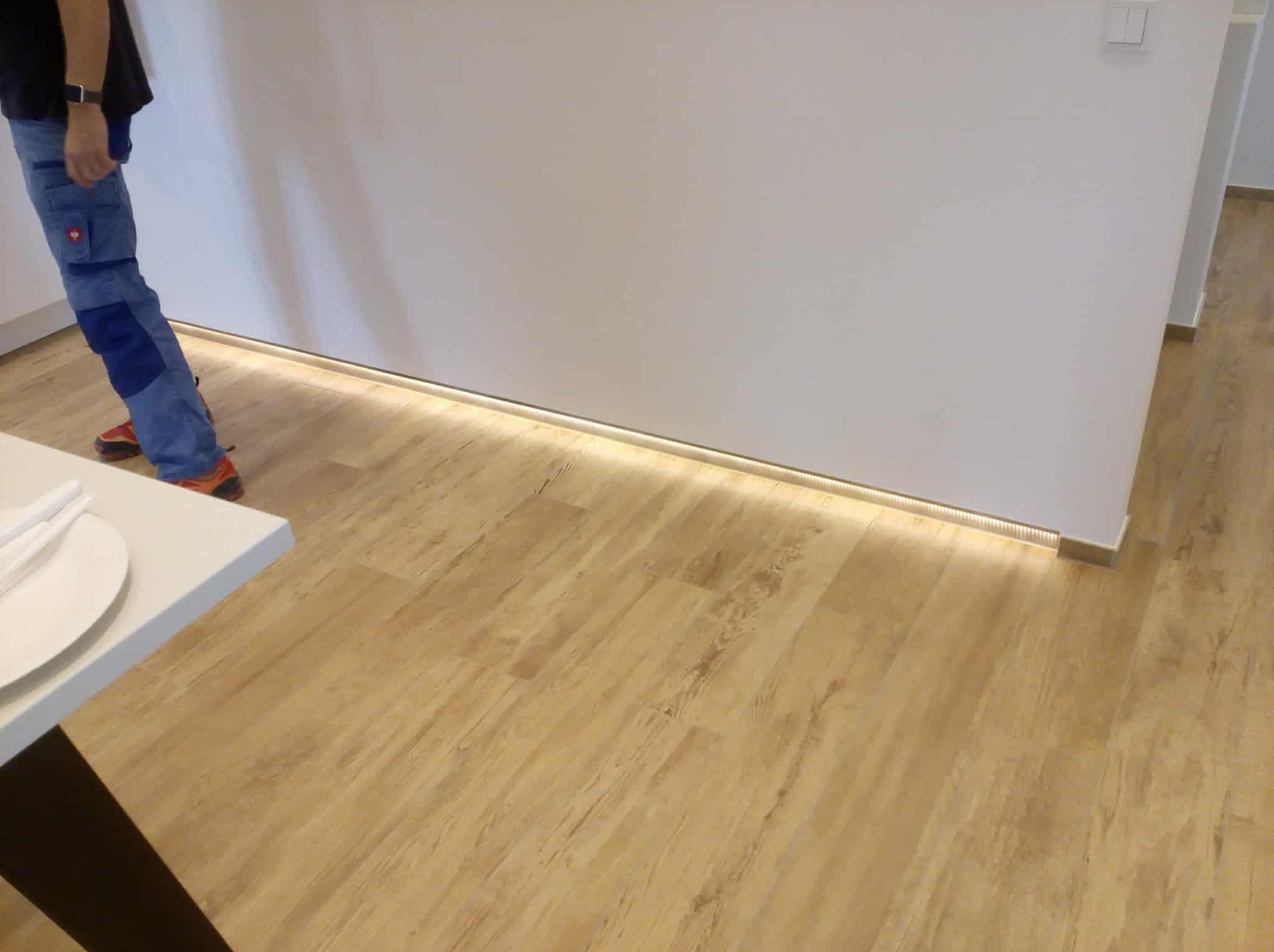 Nowoczesne wnętrze z podświetlaną listwą przypodłogową LED, jasnymi panelami podłogowymi i fragmentem białej ściany. Widoczny fragment osoby w ubraniu roboczym.