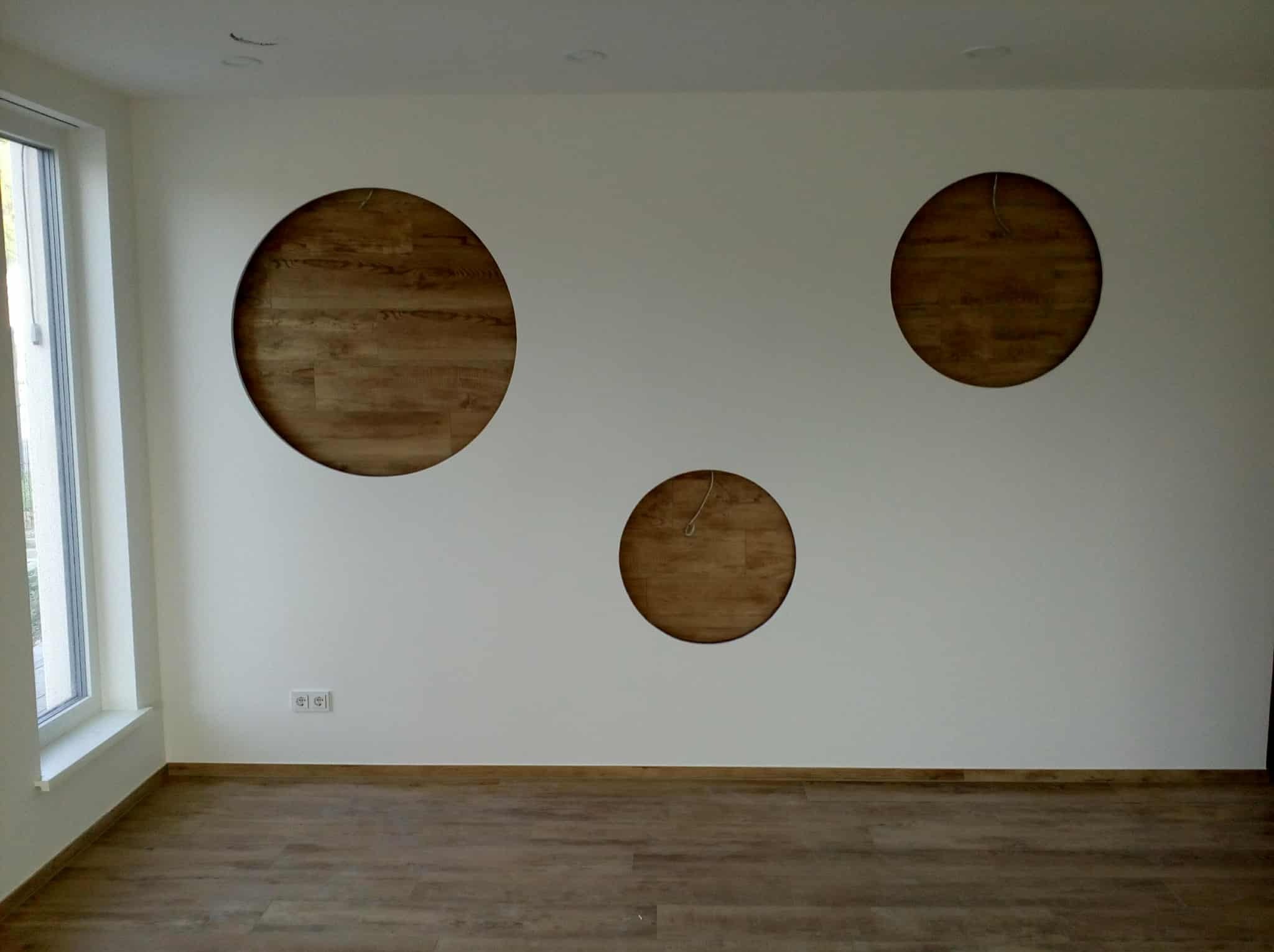 Biała ściana z trzema okrągłymi wnękami wypełnionymi drewnianą okładziną, widok na podłogę z paneli i okno po lewej stronie.
