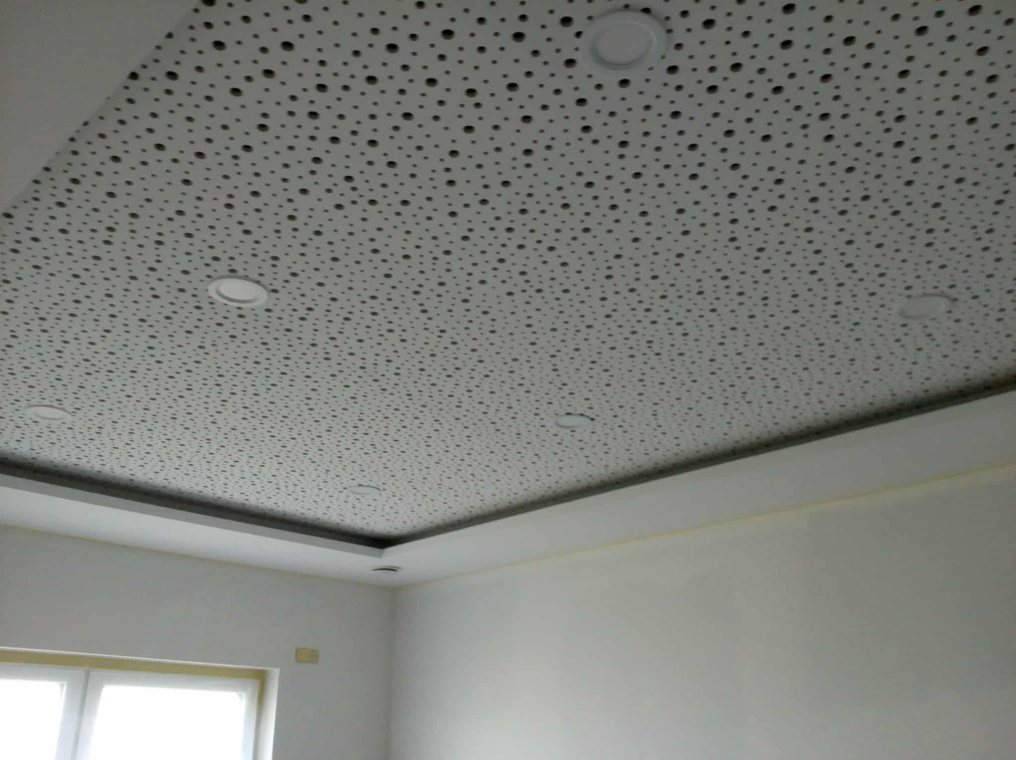 Biały sufit podwieszany z perforacją i wbudowanymi lampami punktowymi w jasnym pomieszczeniu. Widoczne fragmenty ścian i okna.