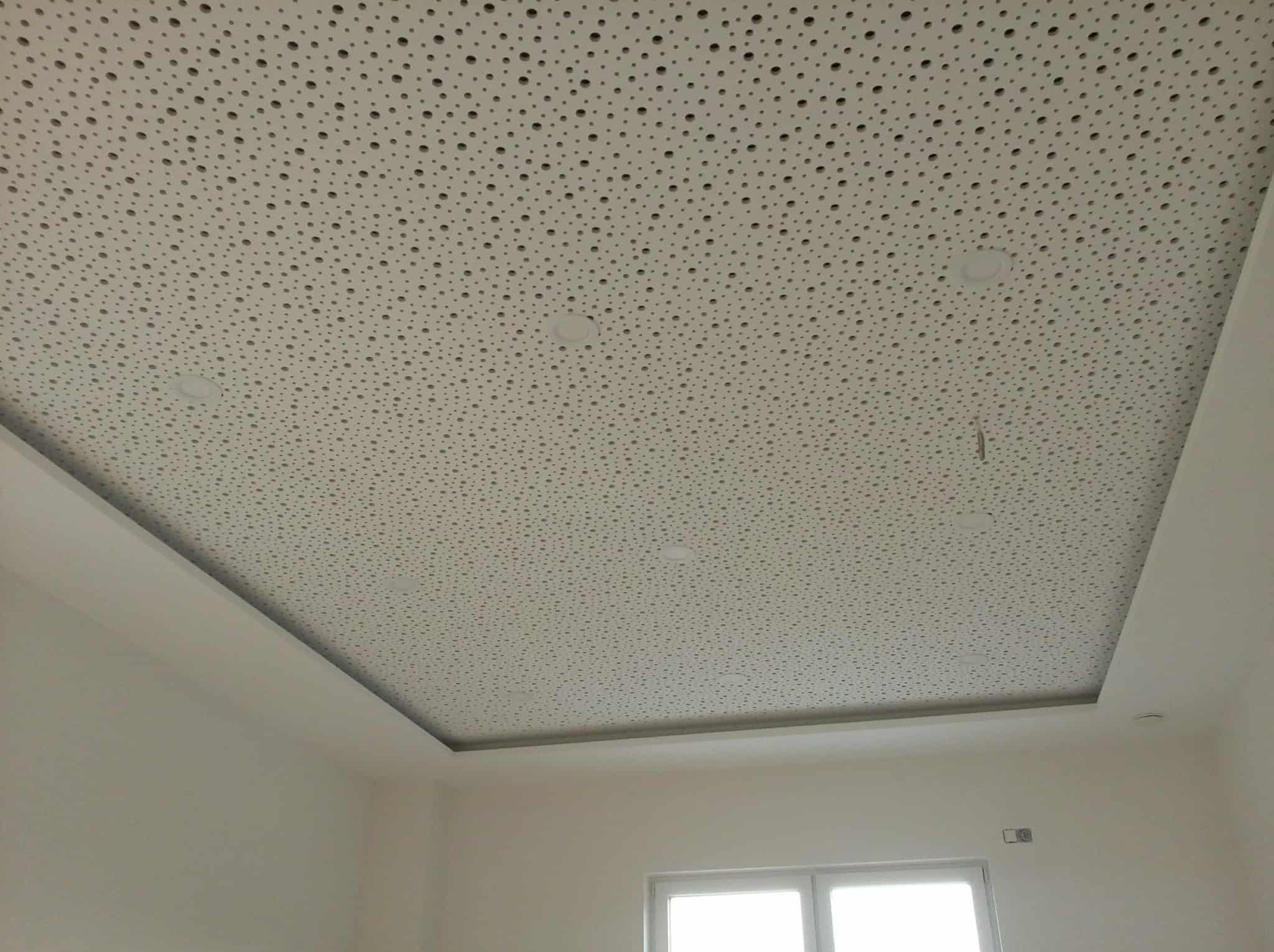 Biały sufit podwieszany z perforacją i wbudowanym oświetleniem punktowym, widok z dołu, fragment ściany i okna w tle.