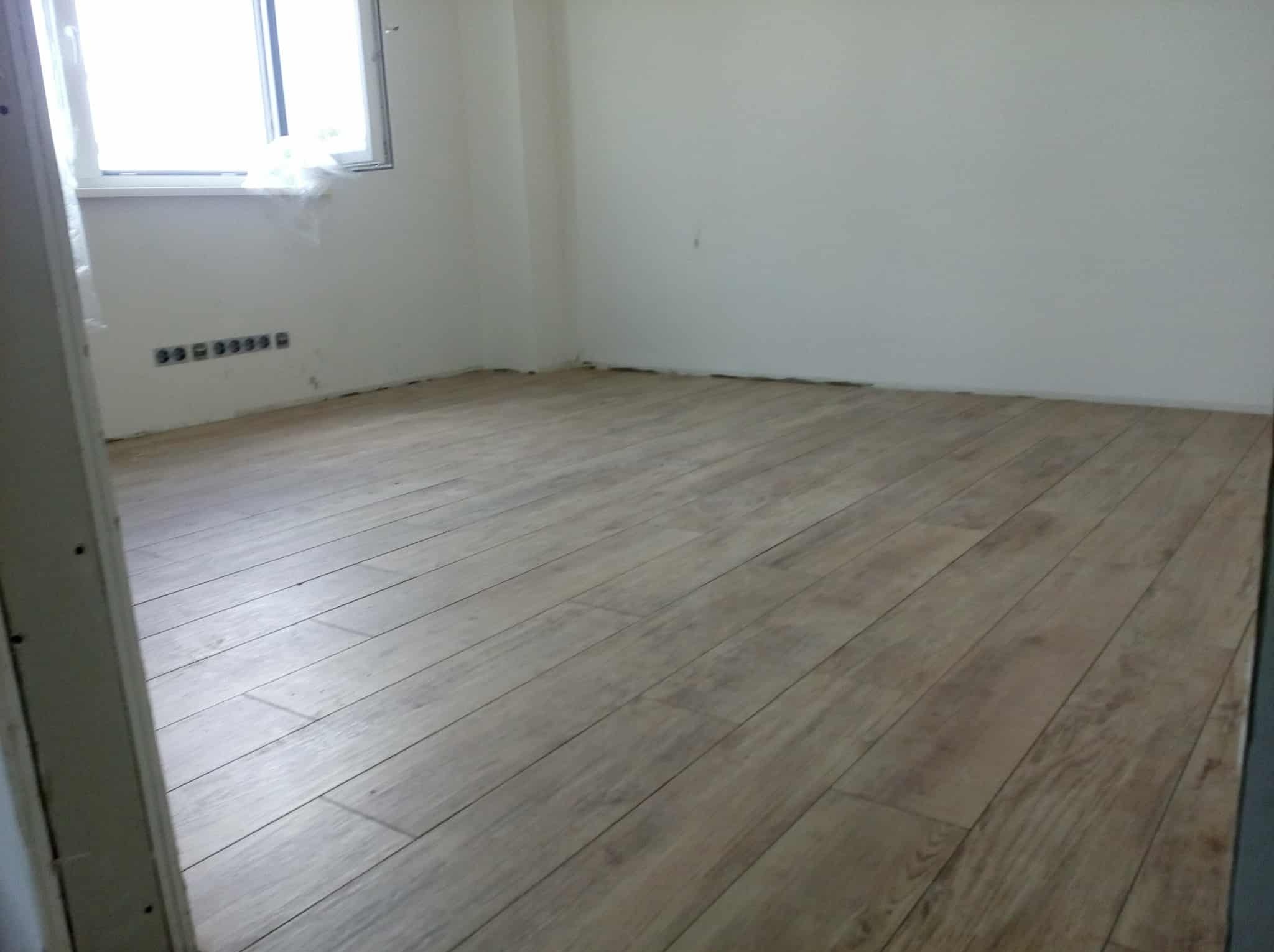 Jasne panele podłogowe ułożone w pustym pokoju z białymi ścianami i oknem. Widoczne gniazdka elektryczne. Ujęcie zza framugi drzwi.