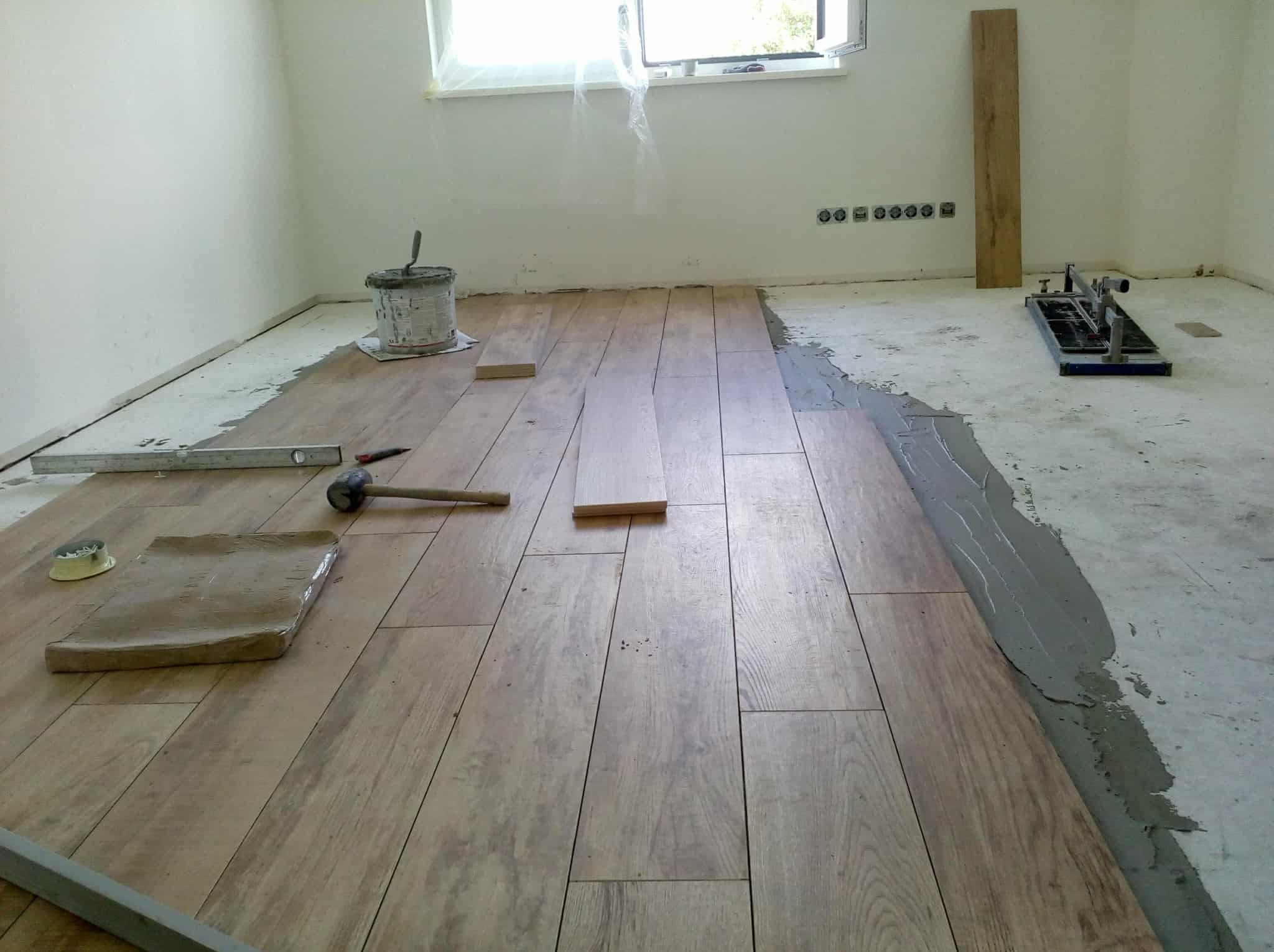 Układanie drewnopodobnych płytek podłogowych w jasnym wnętrzu. Widoczny klej, poziomica, gumowy młotek i maszyna do cięcia płytek. Częściowo ułożona podłoga.