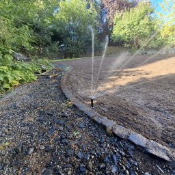 Green Alley Krzysztof Tarasiewicz - Automatyczny system nawadniania w ogrodzie: zraszacze w akcji, nawilżają przygotowaną glebę pod trawnik, obok kamienny chodnik i zieleń. Widok z poziomu gruntu.