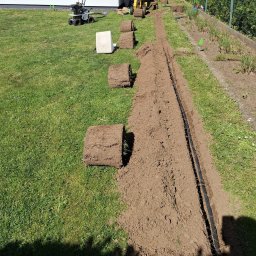 Green Alley Krzysztof Tarasiewicz - Układanie trawy z rolki: przygotowany rów z systemem nawadniania, obok zwinięte rolki trawy i maszyna do rozkładania. Słoneczny dzień, widok z góry.