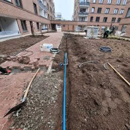 Green Alley Krzysztof Tarasiewicz - Instalacja nawadniania w trakcie budowy ogrodu: wykopane rowy, rury, kostka brukowa, ziemia, budynki w tle. Prace ogrodnicze w toku.