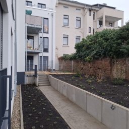 Green Alley Krzysztof Tarasiewicz - Nowoczesny ogród miejski z betonowymi murkami oporowymi, świeżo posadzoną roślinnością i brukowanym chodnikiem prowadzącym do schodów.