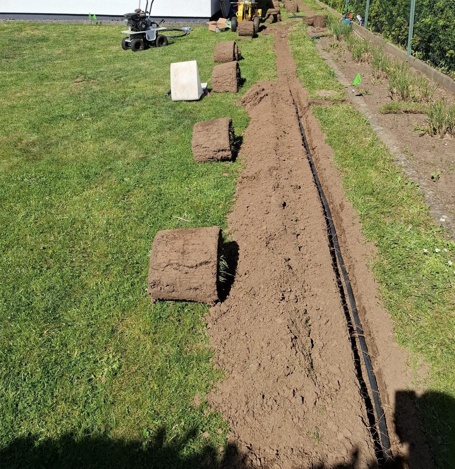 Układanie trawy z rolki: przygotowany rów z systemem nawadniania, obok zwinięte rolki trawy i maszyna do rozkładania. Słoneczny dzień, widok z góry.