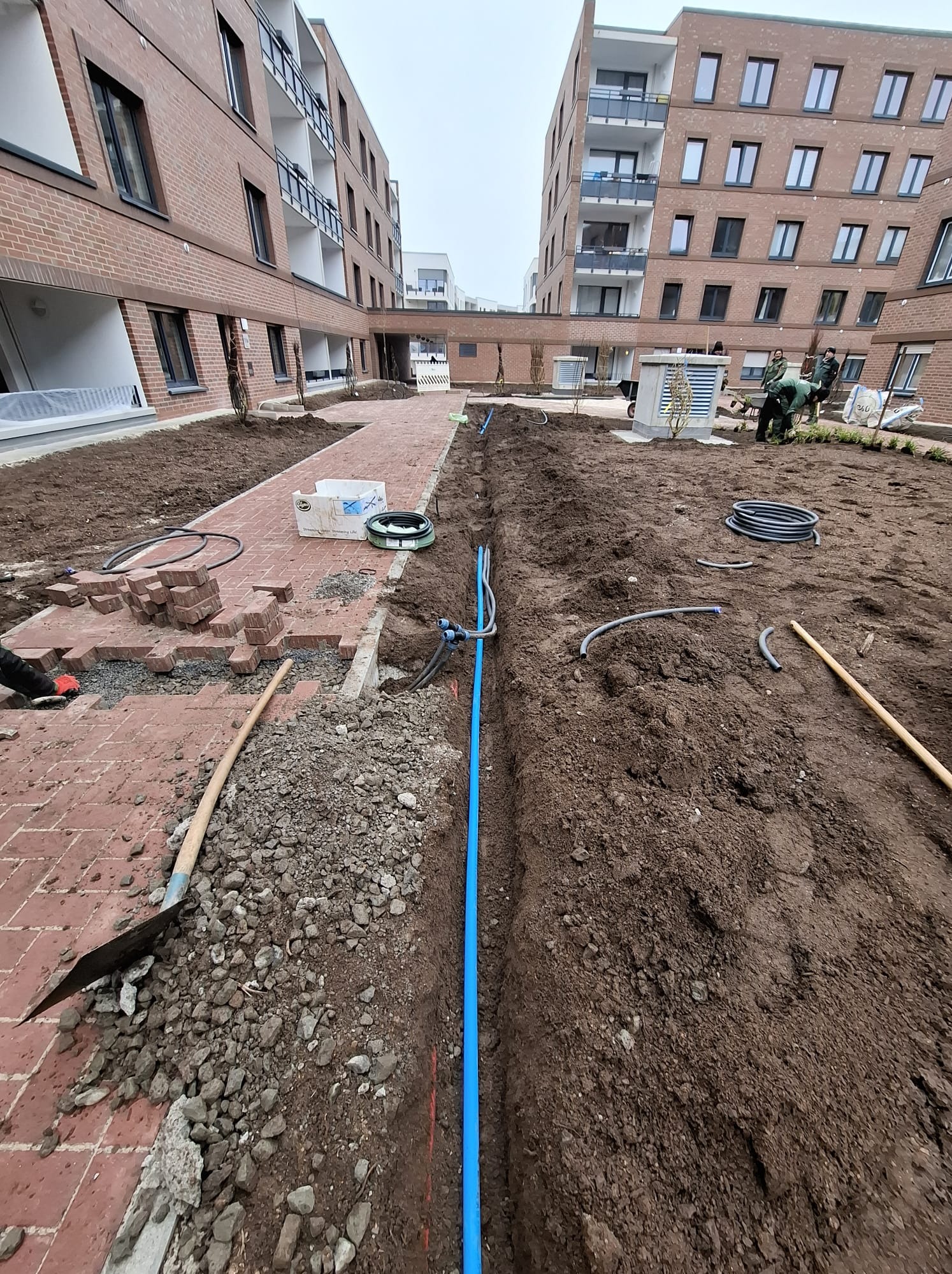 Instalacja nawadniania w trakcie budowy ogrodu: wykopane rowy, rury, kostka brukowa, ziemia, budynki w tle. Prace ogrodnicze w toku.