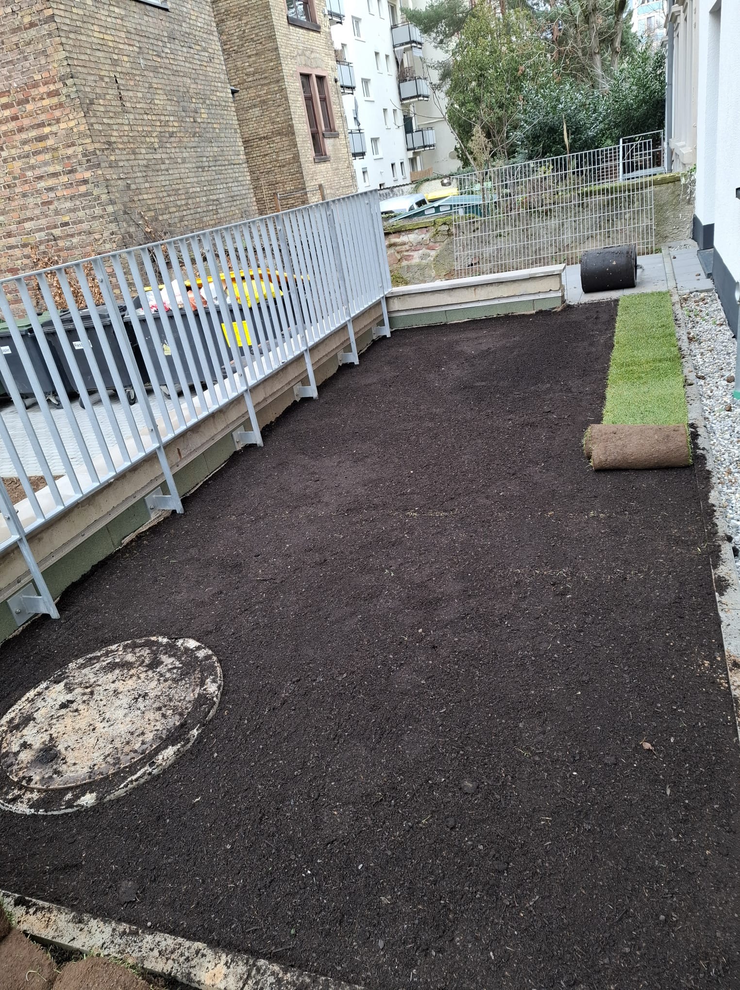 Zakładanie trawnika z rolki na dachu budynku. Częściowo rozłożona trawa, widoczna czarna ziemia i zwinięta rolka. Metalowa barierka w tle.