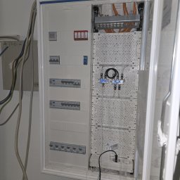 Marcin Kacperski EMAvolt - Otwarta, biała skrzynka elektryczna z bezpiecznikami, rozdzielaczami i przewodami. Widoczne zabezpieczenia przeciwprzepięciowe i gniazdko z wtyczką. Kable na ścianie.