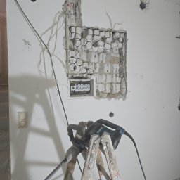 Marcin Kacperski EMAvolt - Rozkuta ściana z widoczną skrzynką elektryczną, kable i bruzdownica na drabinie. Etap prac instalacyjnych, widoczne ślady po kuciu. Przewody elektryczne wystają ze ściany.