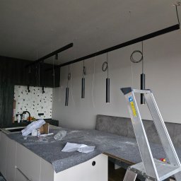Marcin Kacperski EMAvolt - Montaż czarnych lamp cylindrycznych na szynie w kuchni w Słupsku. Widoczna drabina i narzędzia, co sugeruje trwający proces instalacji oświetlenia.
