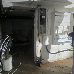 Marcin Kacperski EMAvolt - Rozdzielnia elektryczna z licznikiem i zabezpieczeniami w piwnicy, widoczne kable i instalacje. Osoba w trakcie pracy przy rozdzielnicy.