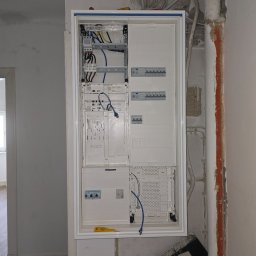 Marcin Kacperski EMAvolt - Rozdzielnica elektryczna w białej obudowie, widoczne kable, bezpieczniki i narzędzie (żółty śrubokręt). Ściana w trakcie remontu, widoczne rury i ślady po kuciu.
