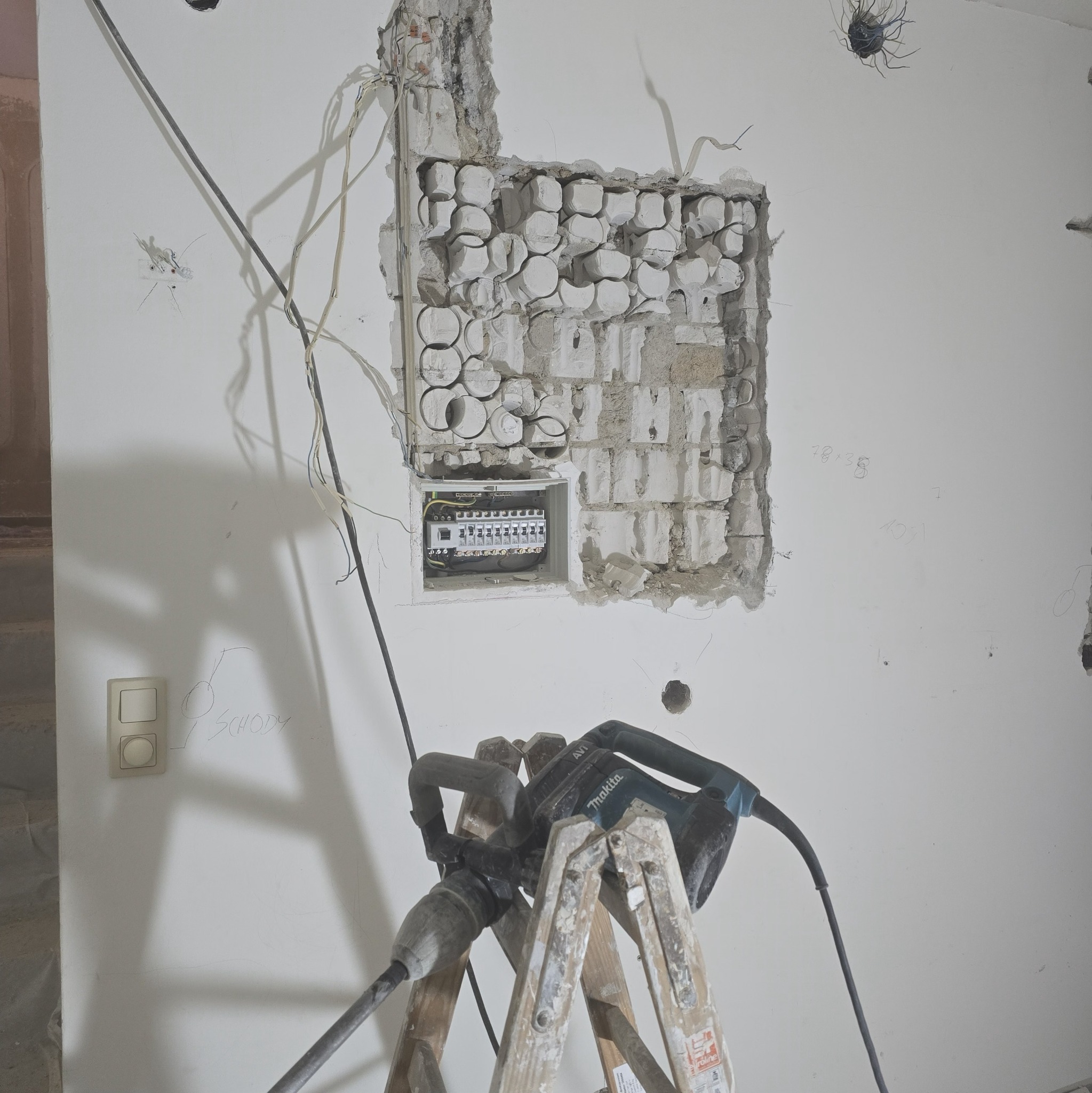 Rozkuta ściana z widoczną skrzynką elektryczną, kable i bruzdownica na drabinie. Etap prac instalacyjnych, widoczne ślady po kuciu. Przewody elektryczne wystają ze ściany.