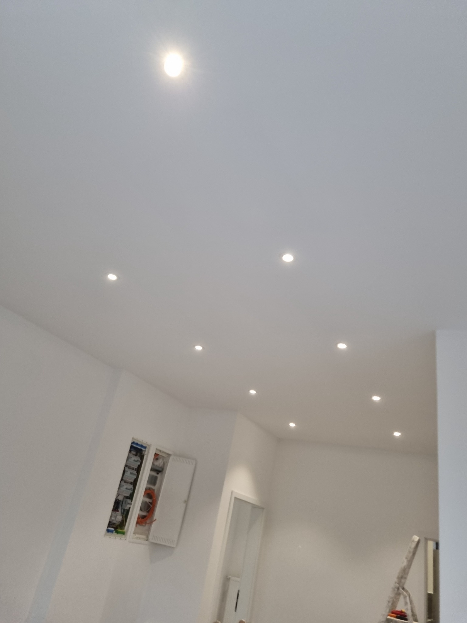 Białe wnętrze z równomiernie rozmieszczonymi punktowymi źródłami światła w suficie. Widoczna otwarta skrzynka elektryczna i fragment drabiny malarskiej.
