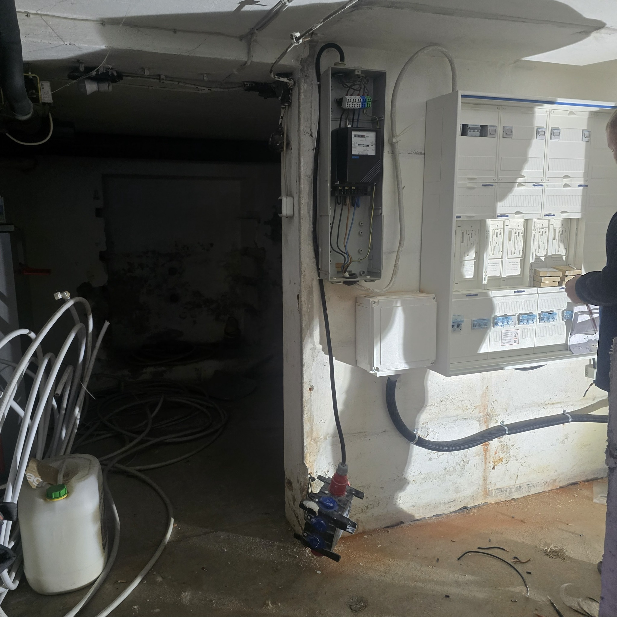 Rozdzielnia elektryczna z licznikiem i zabezpieczeniami w piwnicy, widoczne kable i instalacje. Osoba w trakcie pracy przy rozdzielnicy.