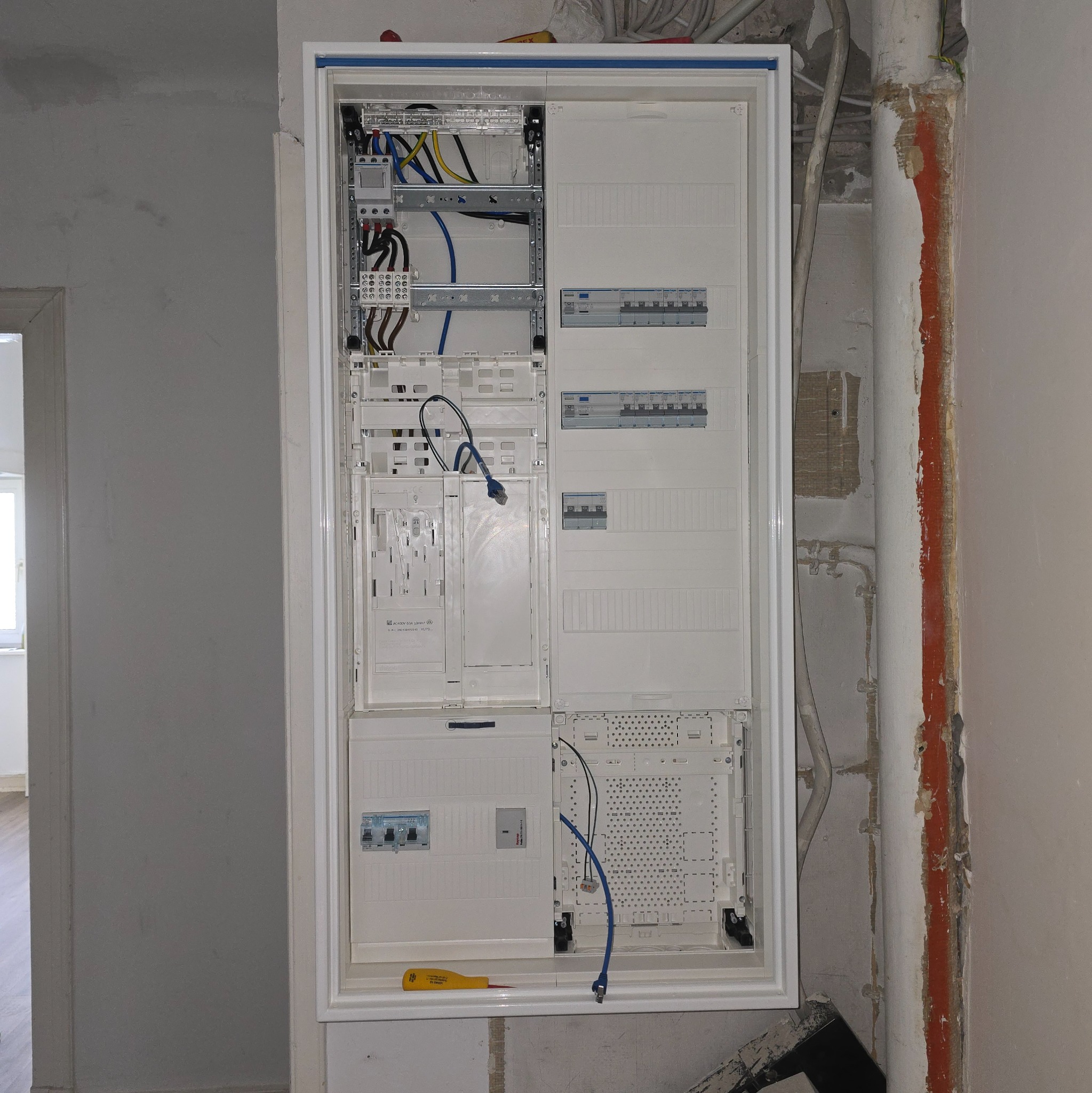 Rozdzielnica elektryczna w białej obudowie, widoczne kable, bezpieczniki i narzędzie (żółty śrubokręt). Ściana w trakcie remontu, widoczne rury i ślady po kuciu.