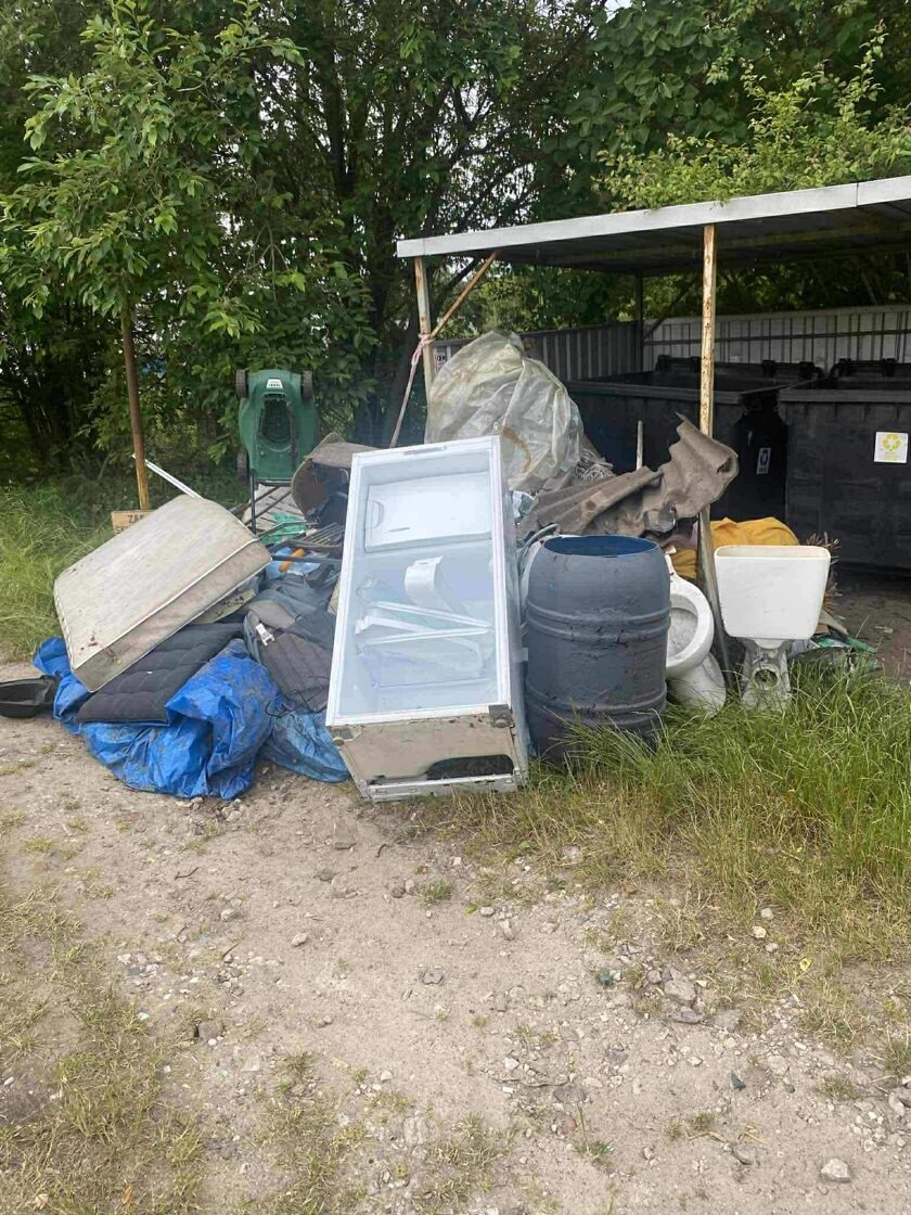Sterta odpadów: stara lodówka z otwartymi drzwiami, materac, beczka, umywalka i inne śmieci na zewnątrz, z zielenią w tle. Widoczne kontenery na odpady.