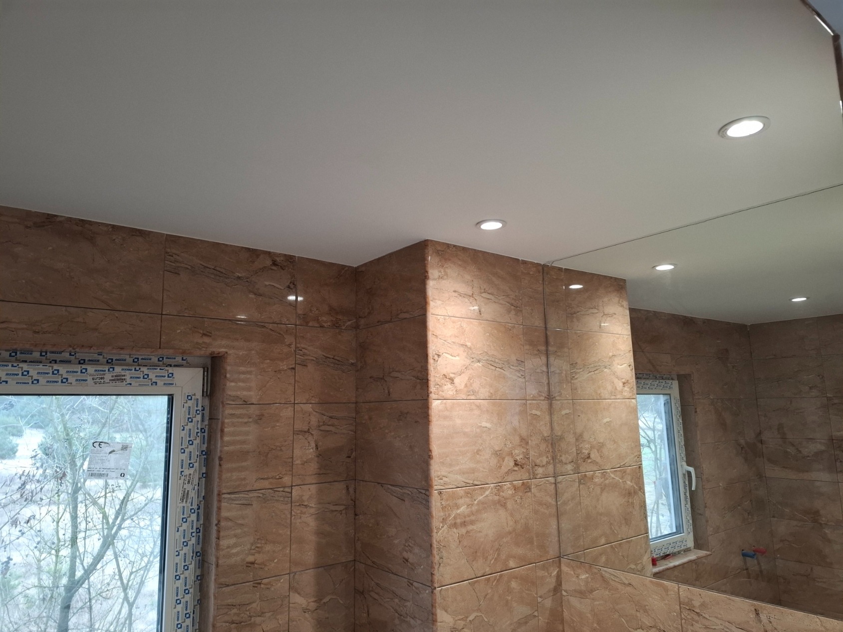 Wykończona łazienka z beżowymi płytkami imitującymi marmur, podwieszanym sufitem z oświetleniem punktowym i oknem z widokiem na drzewa.