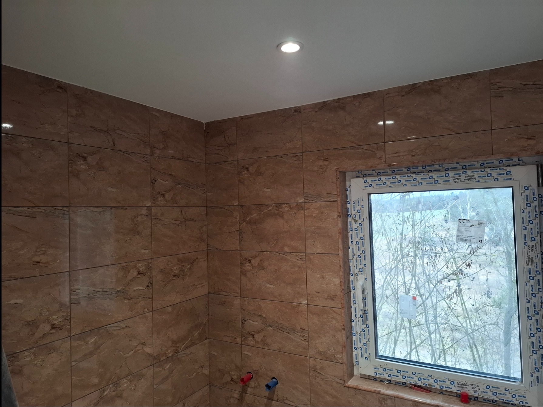 Wykończona łazienka z brązowymi płytkami imitującymi marmur, białe okno z widokiem na drzewa, sufit z oświetleniem punktowym. Widoczne przyłącza wodne.