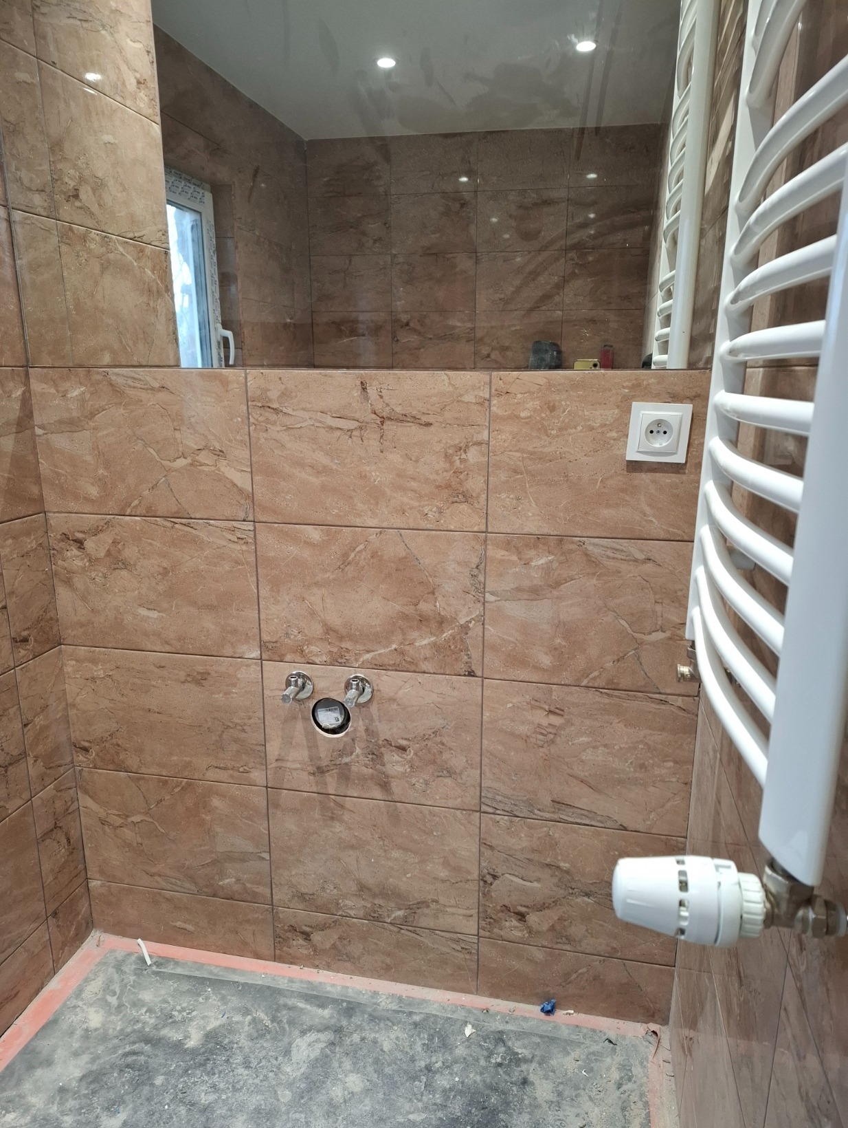 Wykończona łazienka z brązowymi płytkami imitującymi marmur, widoczny grzejnik drabinkowy i przygotowane przyłącze do baterii.