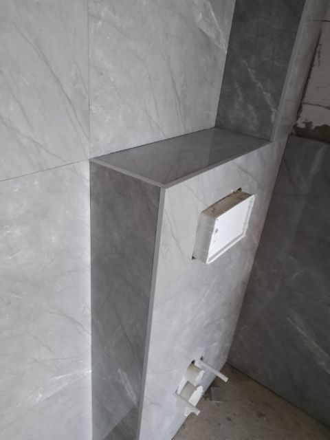 Wykończona wnęka z półką i ukrytą instalacją wodną, obłożona szarymi płytkami imitującymi marmur. Minimalistyczny design, precyzyjne ułożenie płytek.