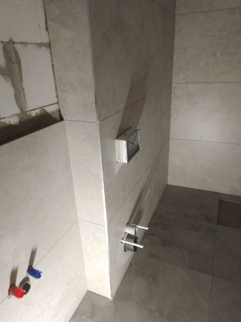 Wykończona łazienka z beżowymi płytkami imitującymi kamień, widoczne przyłącza wodne i stelaż podtynkowy WC. Minimalistyczny design, nowoczesne rozwiązania.