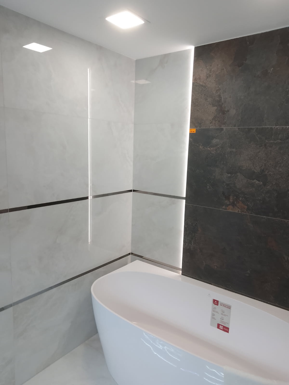 Nowoczesna łazienka z białą wanną, jasnymi płytkami imitującymi marmur i ciemnymi płytkami z efektem kamienia. Oświetlenie LED podkreśla pionowe linie.