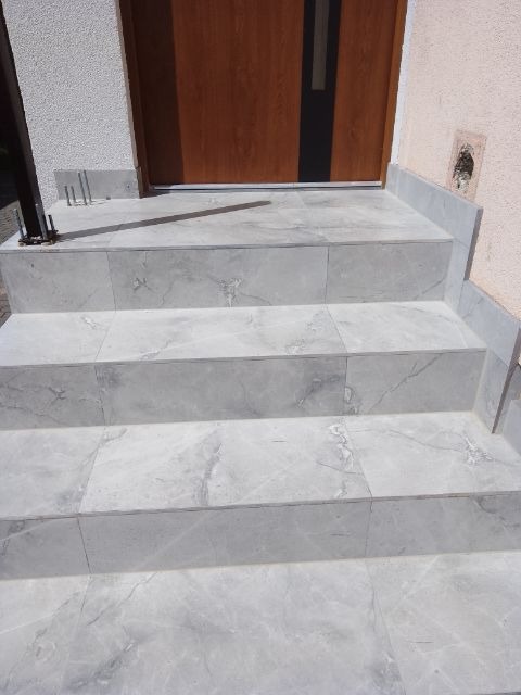 Wykończone marmurowe schody wejściowe do domu z drewnianymi drzwiami. Widoczne detale wykończenia i szare płytki z marmurowym wzorem.