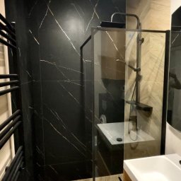 Mykyta Sichkar S-KAR BUD - Wykończona łazienka z czarnymi płytkami imitującymi marmur ze złotymi żyłami, kabiną prysznicową typu walk-in i grzejnikiem drabinkowym w kolorze czarnym.