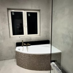 Mykyta Sichkar S-KAR BUD - Narożna wanna obudowana mozaiką w wykończonej łazience. Widoczne złote baterie, okno i szklana kabina prysznicowa. Ściany wykończone w szarym odcieniu.