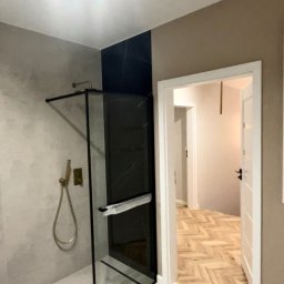 Mykyta Sichkar S-KAR BUD - Nowoczesna łazienka po remoncie: kabina prysznicowa z czarnymi panelami, złota armatura, biały grzejnik drabinkowy i parkiet w jodełkę w tle.