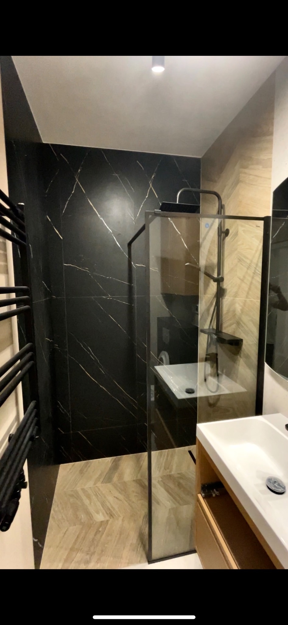 Wykończona łazienka z czarnymi płytkami imitującymi marmur ze złotymi żyłami, kabiną prysznicową typu walk-in i grzejnikiem drabinkowym w kolorze czarnym.