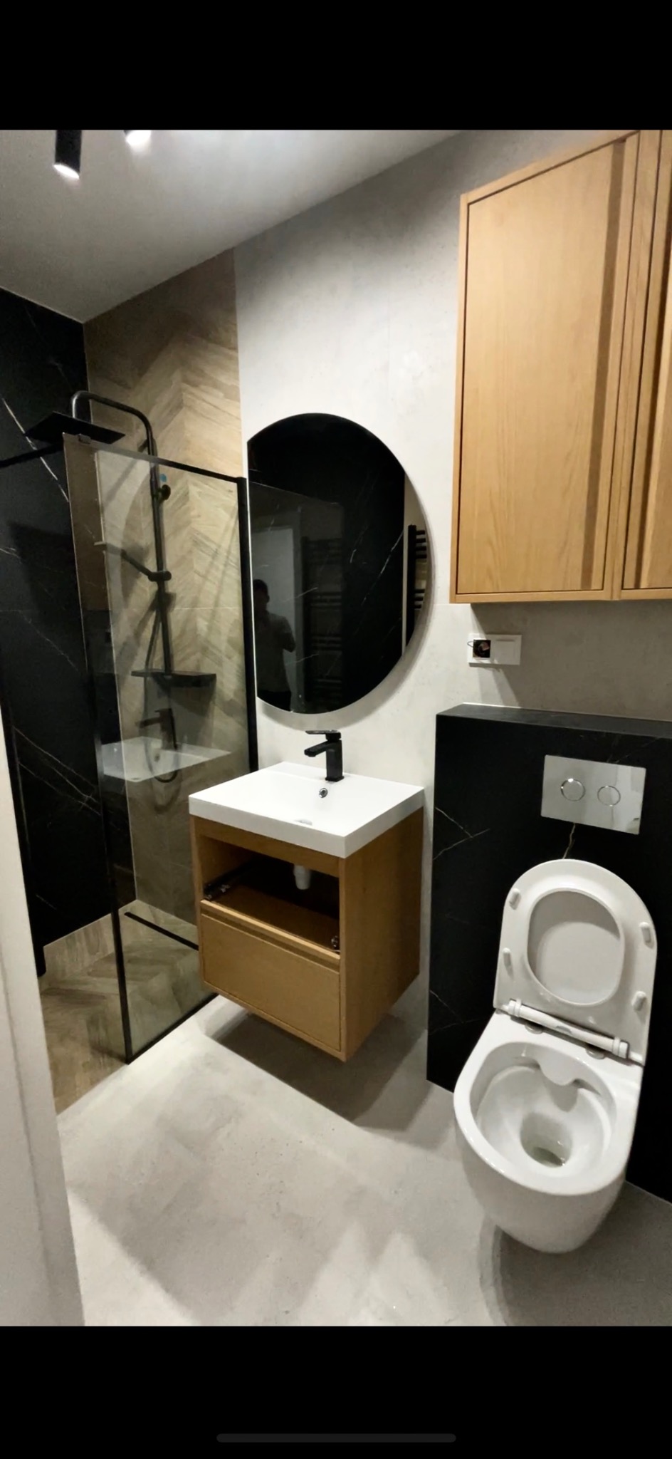 Nowoczesna łazienka z kabiną prysznicową typu walk-in, okrągłym lustrem i wiszącą szafką z umywalką. Uwagę zwraca czarna ściana z białymi żyłami.