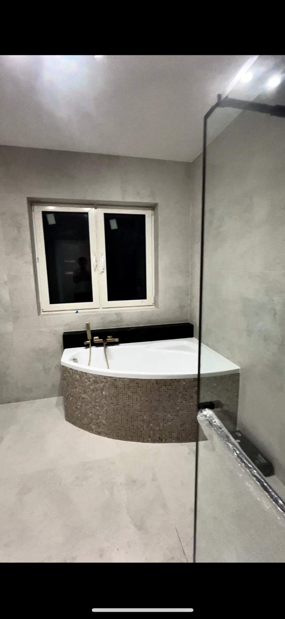 Narożna wanna obudowana mozaiką w wykończonej łazience. Widoczne złote baterie, okno i szklana kabina prysznicowa. Ściany wykończone w szarym odcieniu.