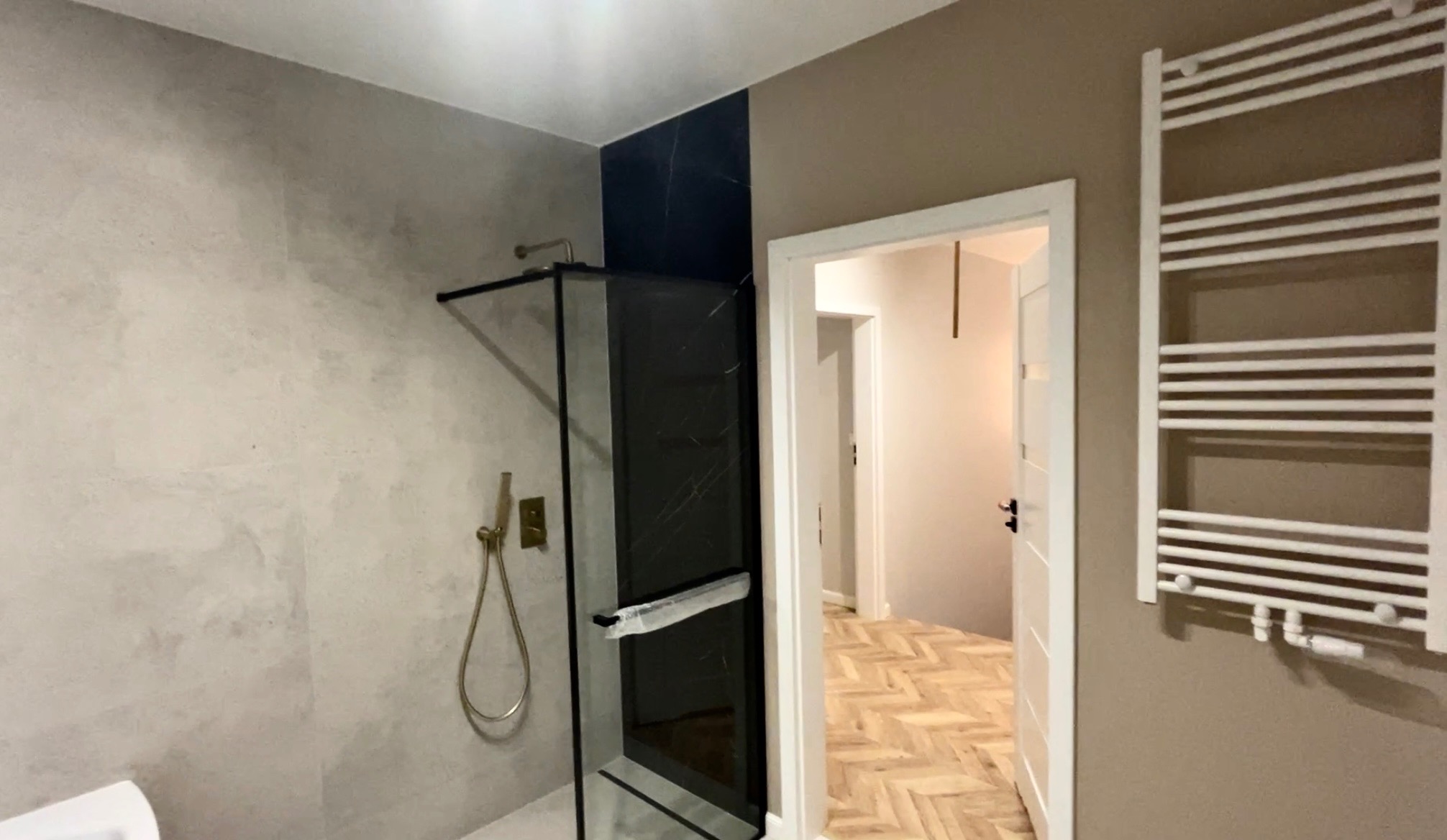 Nowoczesna łazienka po remoncie: kabina prysznicowa z czarnymi panelami, złota armatura, biały grzejnik drabinkowy i parkiet w jodełkę w tle.