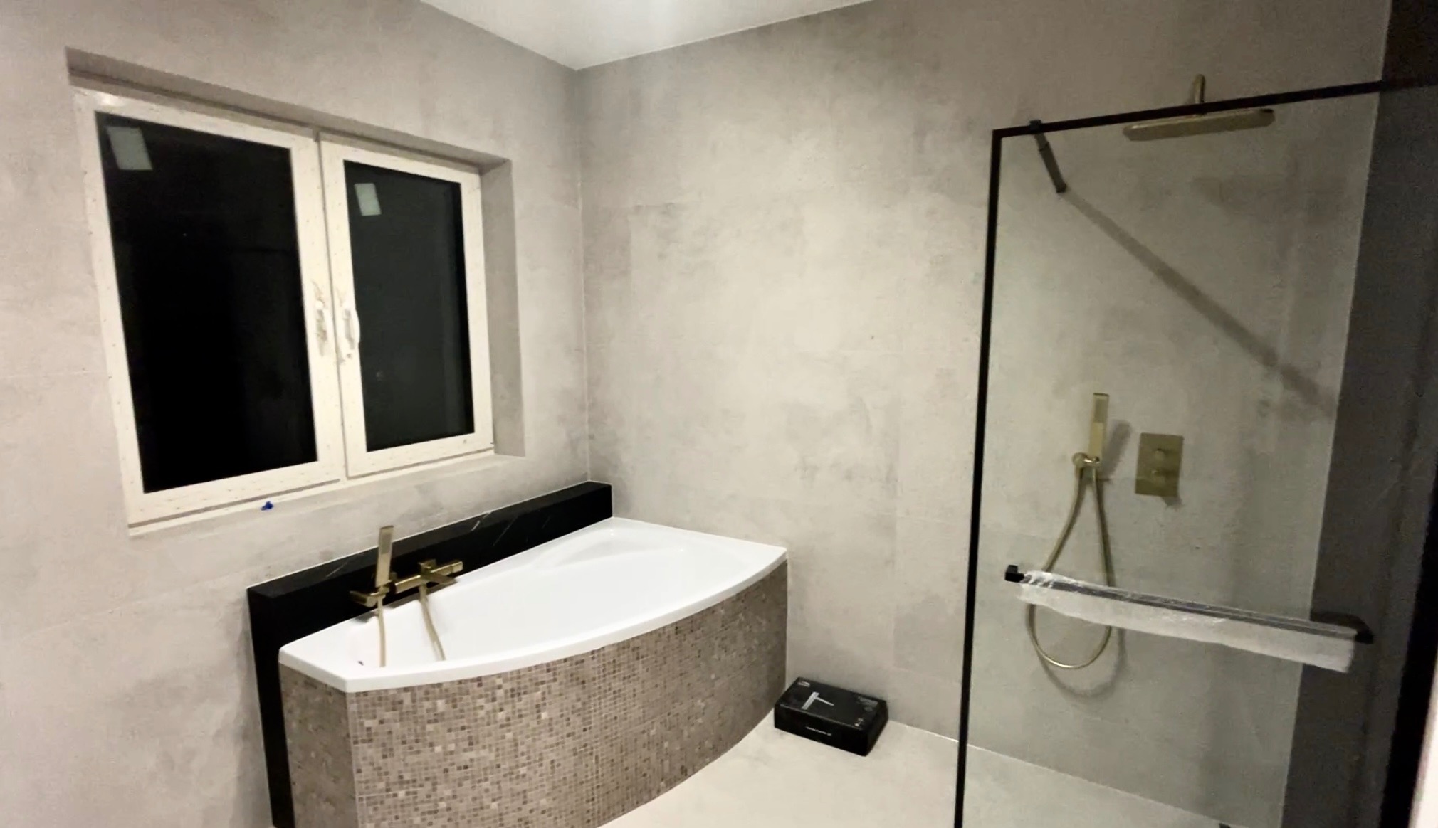 Nowoczesna łazienka z wanną obudowaną mozaiką, czarnym blatem i złotą armaturą. Kabina prysznicowa ze złotym zestawem natryskowym. Szare płytki na ścianach i białe okno.