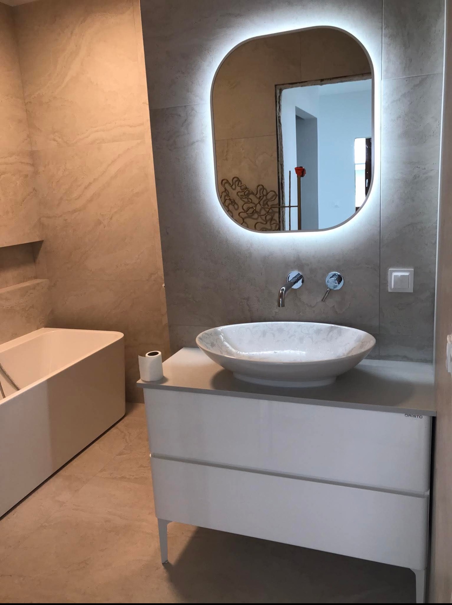 Wykończona łazienka z beżowymi płytkami, białą wanną i umywalką nablatową. Nad umywalką podświetlane lustro o zaokrąglonym kształcie. Widoczny fragment toalety.