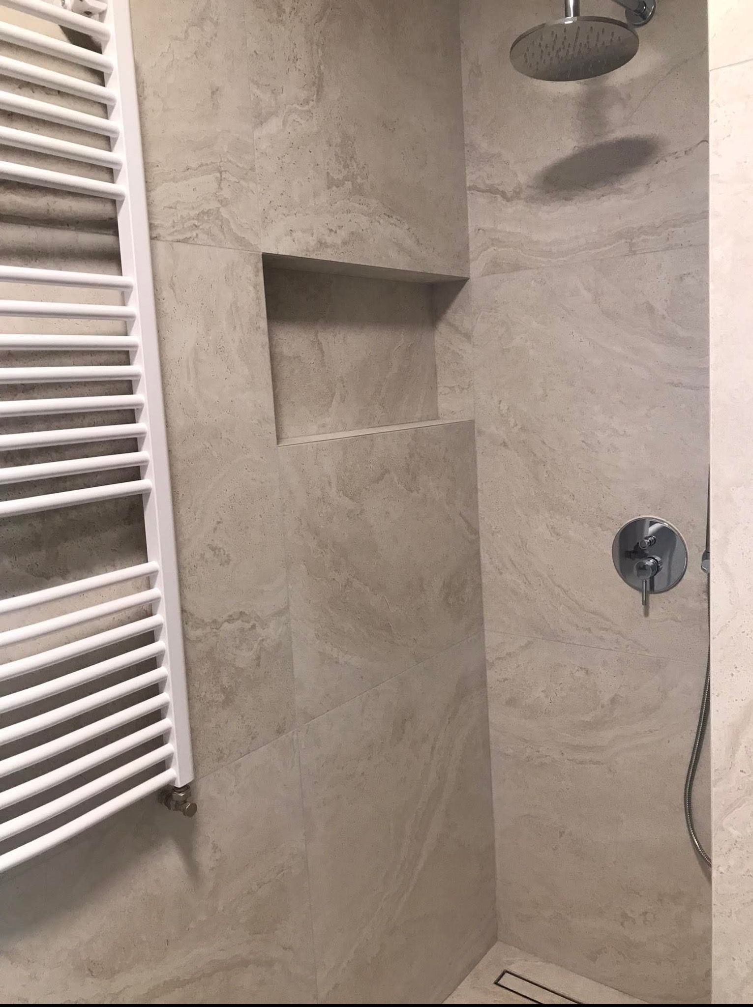 Wykończona kabina prysznicowa z beżowymi płytkami imitującymi kamień, wnęką i grzejnikiem drabinkowym. Nowoczesna armatura i odpływ liniowy.