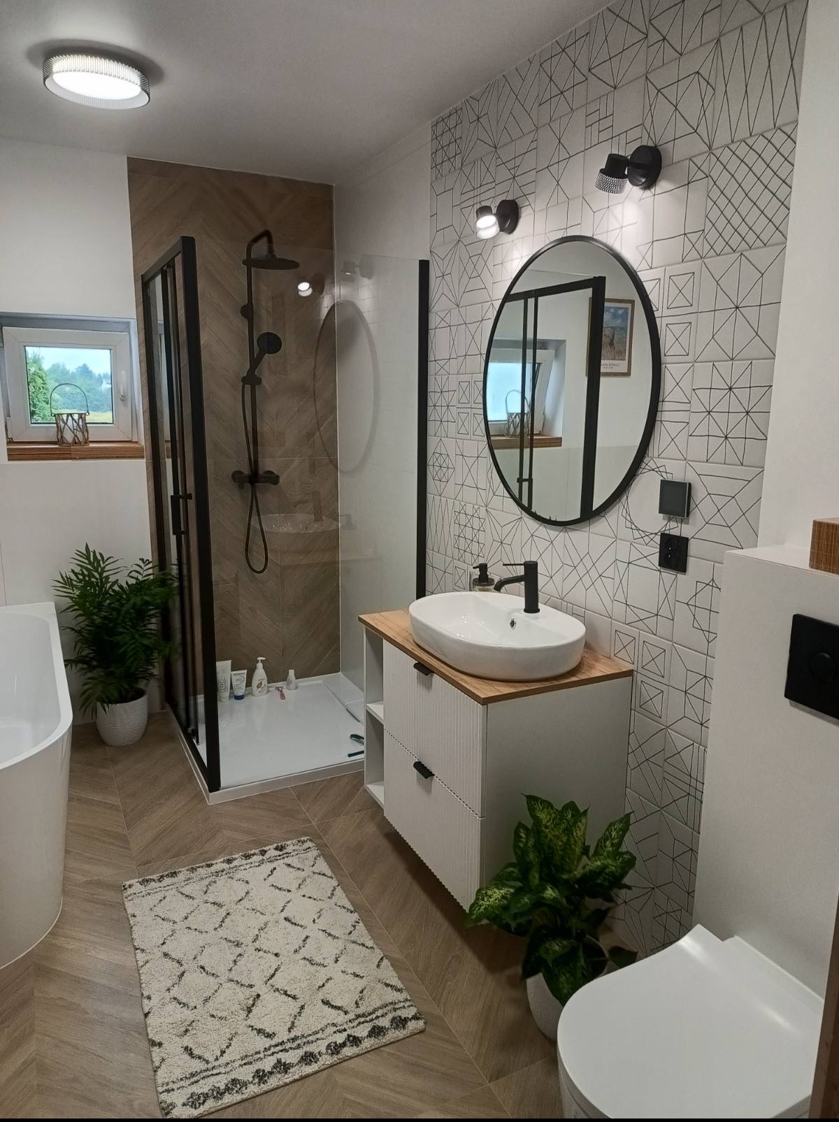 Nowoczesna łazienka z kabiną prysznicową typu walk-in, umywalką nablatową na szafce z drewnianym blatem i geometrycznymi płytkami na ścianie.