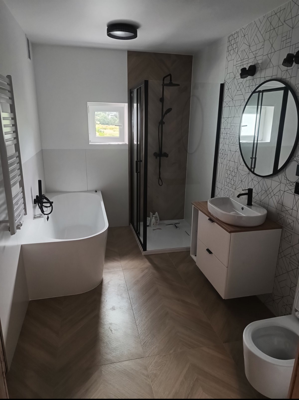 Nowoczesna łazienka po remoncie: biała wanna, kabina prysznicowa z czarnymi ramami, umywalka na szafce z drewnianym blatem, lustro w czarnej ramie i wzorzyste płytki.
