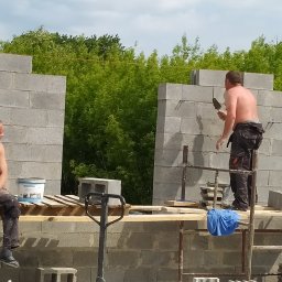 UKR-POL BUD Yurii Herchuk - Wznoszenie ścian z bloczków betonowych na budowie domu jednorodzinnego. Dwóch robotników na rusztowaniu, jeden muruje, drugi odpoczywa. Widok na zieleń w tle.